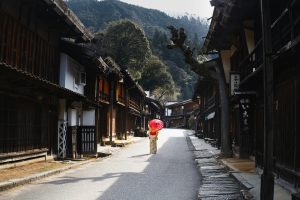 Scenic Japanese village street with a woman in kimono carrying a red umbrella, capturing cultural essence.