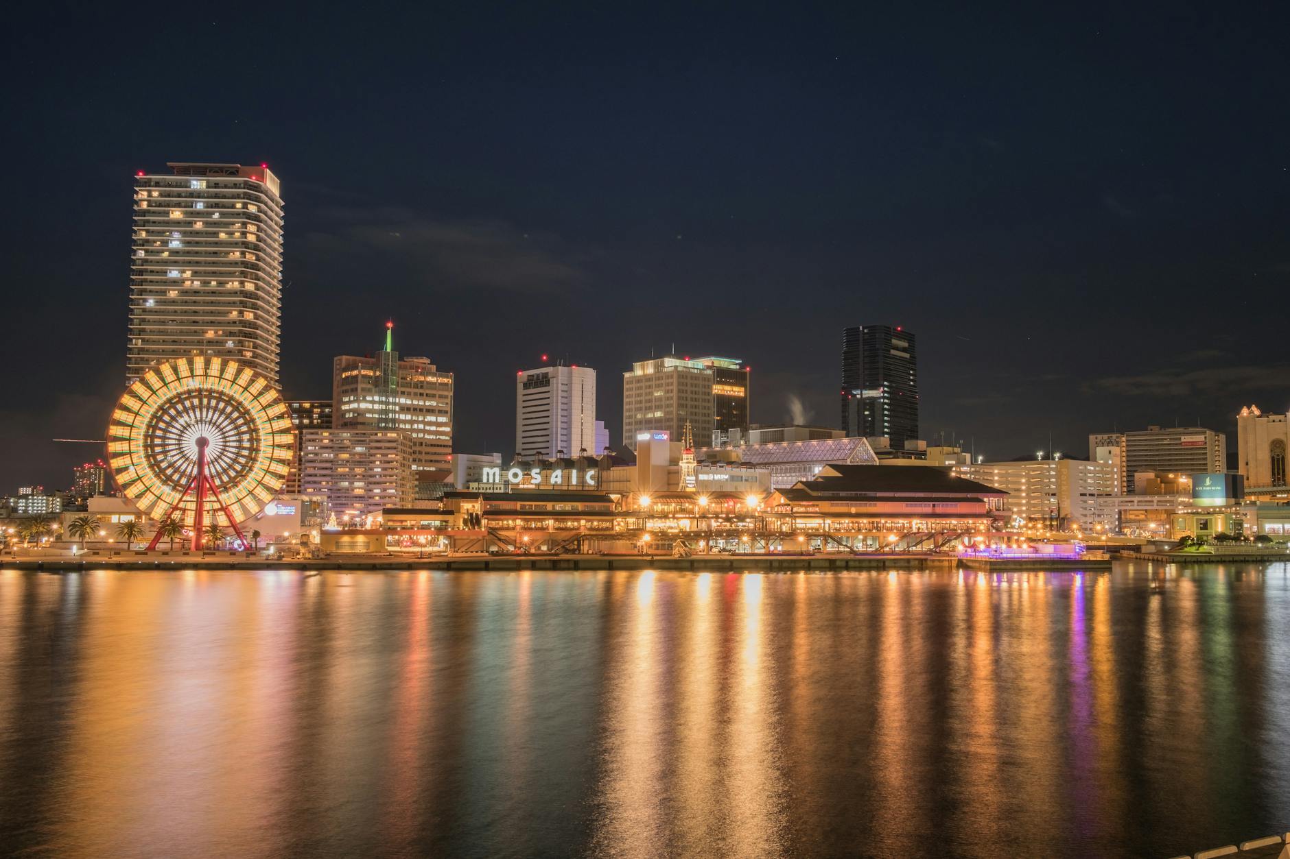 Stunning night cityscape of Kobe with illuminated Ferris wheel reflecting on the waterfront.